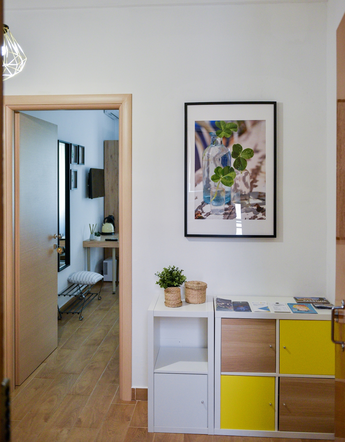 A modern corridor is observed, featuring light-colored walls and wooden flooring. A framed artwork displaying a bottle with clovers is hung above a storage unit with yellow and wood-tone drawers. A door leads to a cozy room, with light spilling in from the entrance.
