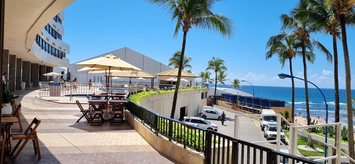 A sunlit outdoor terrace is featured, lined with palm trees and equipped with wooden tables and umbrellas. The ocean can be seen in the background, with the blue sky above creating a bright, inviting environment.
