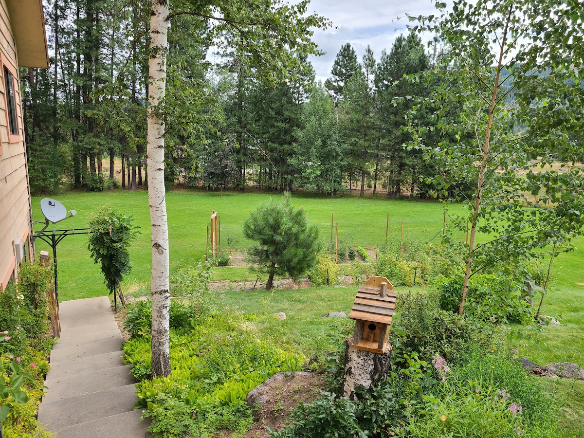 A well-maintained garden features lush green grass and neatly arranged plant beds. A birdhouse sits on a wooden post, while tall trees line the background. A stone pathway leads down from the house, creating a welcoming entry into this peaceful outdoor space.