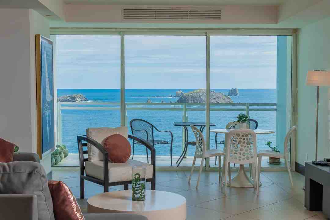 A light-filled living space showcases large glass doors opening to a private balcony with ocean views. A round dining table and several chairs offer seating, while a cozy sitting area is furnished with a soft couch and side tables. Natural coastal scenery is visible.