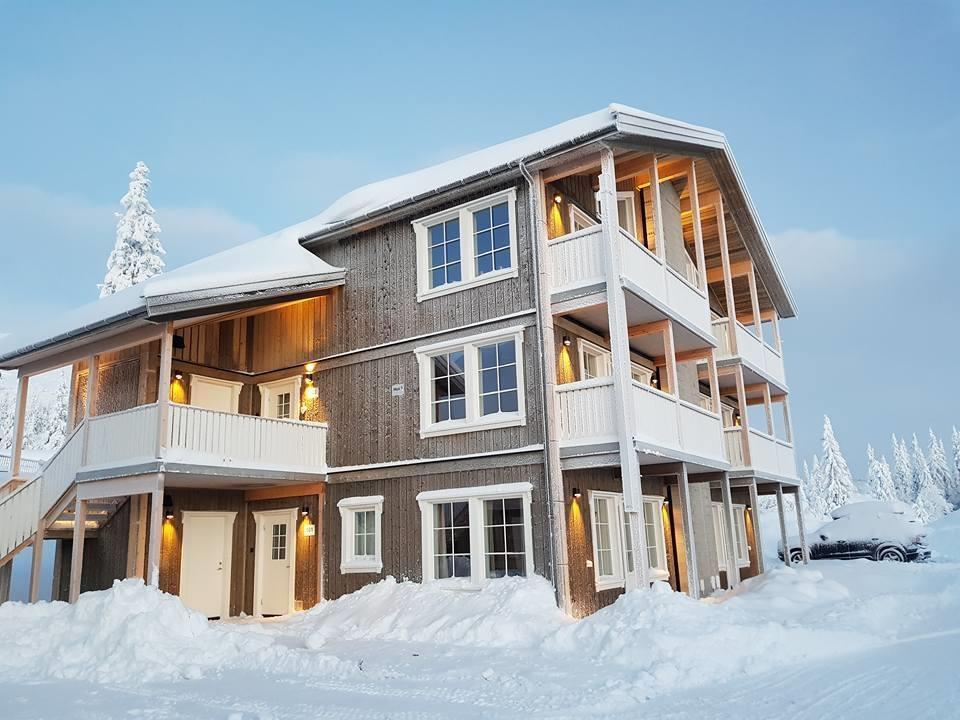 A three-story apartment building is shown, featuring wooden siding and multiple balconies. Snow covers the ground and decorates the entrance pathways. Soft lighting illuminates the exterior, creating a warm ambiance against the snowy landscape. Trees in the background suggest a winter environment.