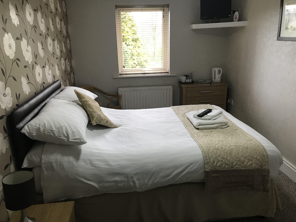 A well-appointed bedroom features a spacious bed adorned with a neutral-toned quilt. A small nightstand holds a lamp, and natural light filters through a window offering a view of greenery. A TV and tea-making facilities are present, with additional furniture arranged for comfort.