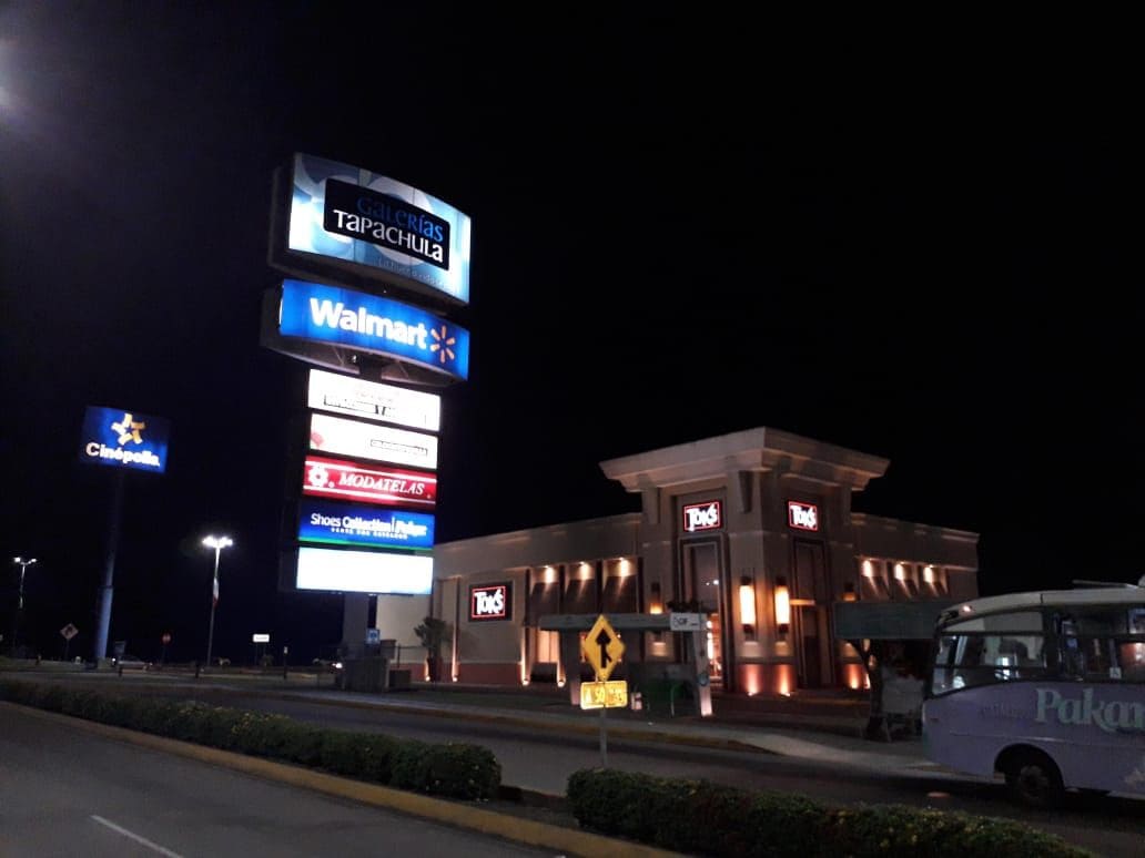 The image depicts a nighttime view of a shopping complex featuring prominent signage for various establishments, including Walmart and Toks. The well-lit area reveals a modern building with a classic design, surrounded by neatly landscaped bushes and a clear road leading to the complex.