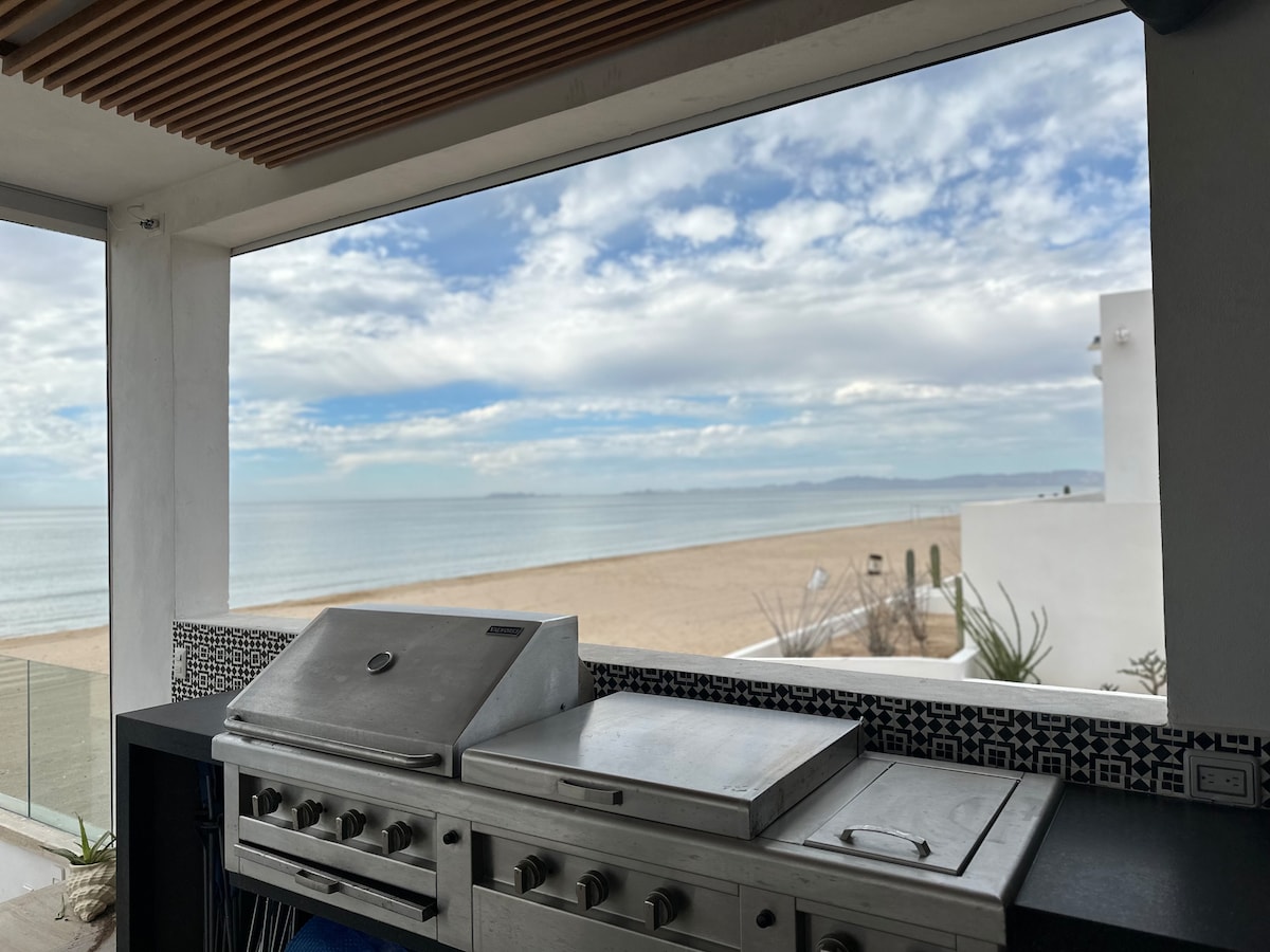 A grilling station is visible, featuring stainless steel equipment set against a backdrop of expansive ocean views. The serene beach stretches out, framed by a large window that enhances the connection between indoor and outdoor spaces.