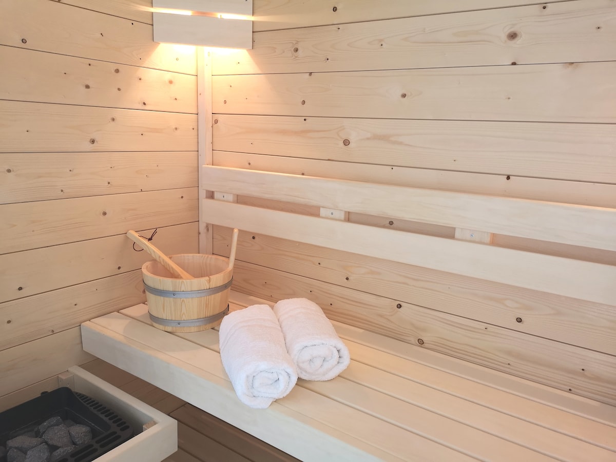 The private sauna features wooden paneling and seating areas. Fresh towels are neatly placed on the bench, and a wooden bucket accompanied by a ladle is visible. A light fixture overhead provides warm illumination, creating a calm and relaxing environment.