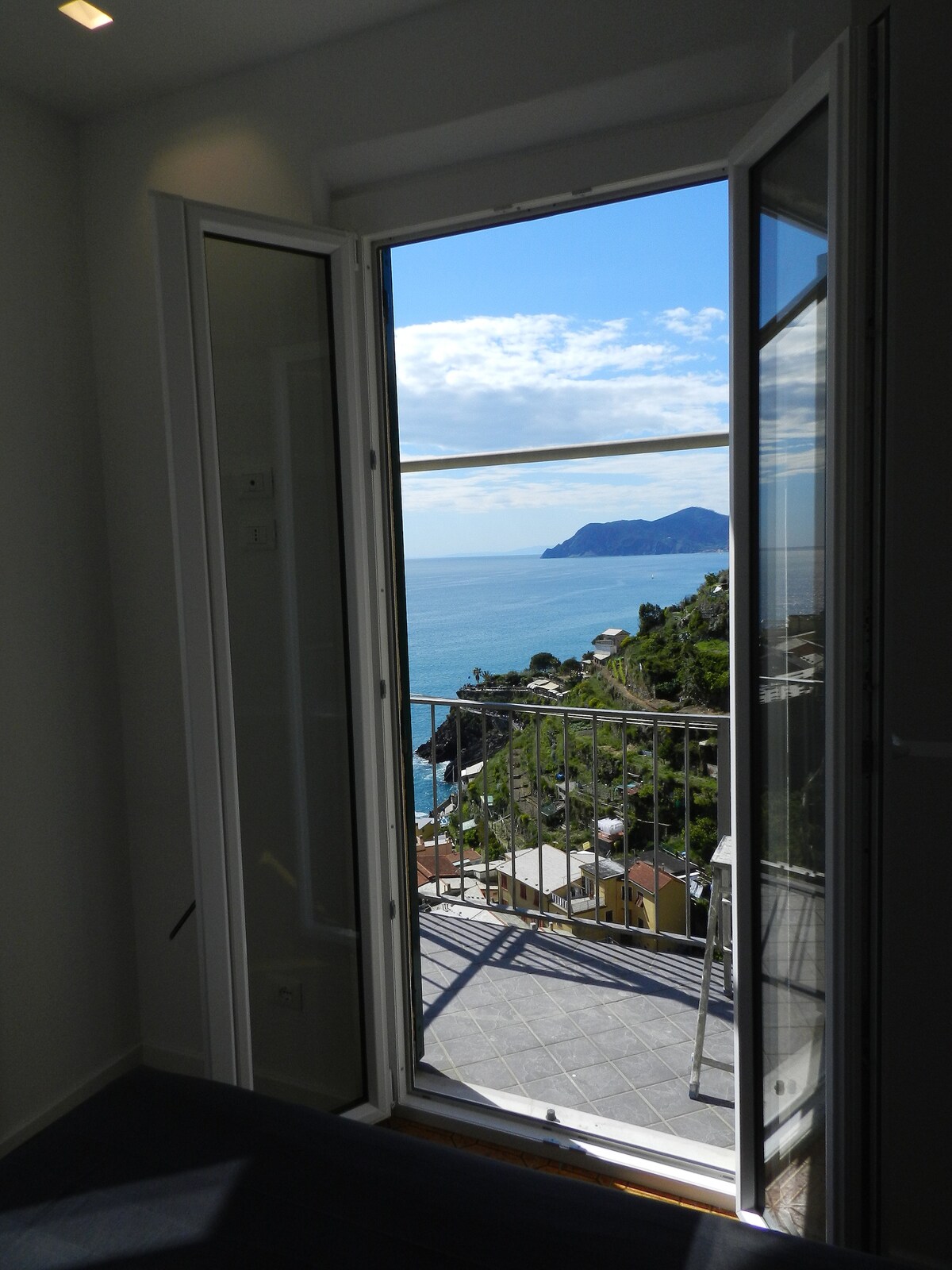 A balcony door stands open, revealing stunning views of the coastline and the sea. The scenic landscape includes distant hills and a few structures nestled along the shoreline. Natural light floods the interior space, enhancing the connection between indoors and outdoors.
