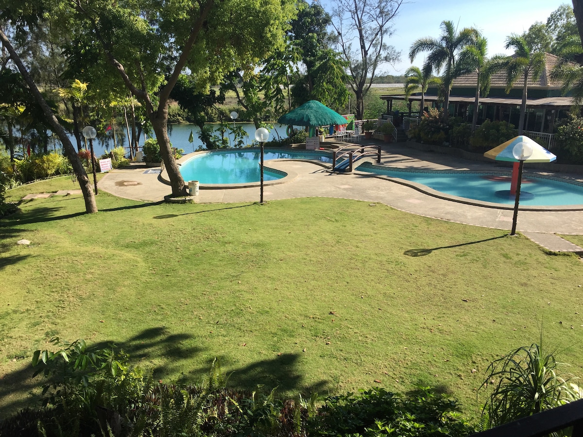 A well-maintained outdoor area features two swimming pools surrounded by lush greenery. Palm trees and vibrant foliage frame the scene, while sun loungers and umbrellas are positioned nearby. The serene view overlooking a river adds a peaceful ambiance to the space.