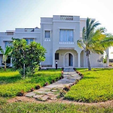 A modern two-story house is set against a clear sky, featuring large windows and elegant architectural details. A pathway made of stone leads through the well-maintained green lawn, with palm trees and shrubs adding to the inviting landscape.
