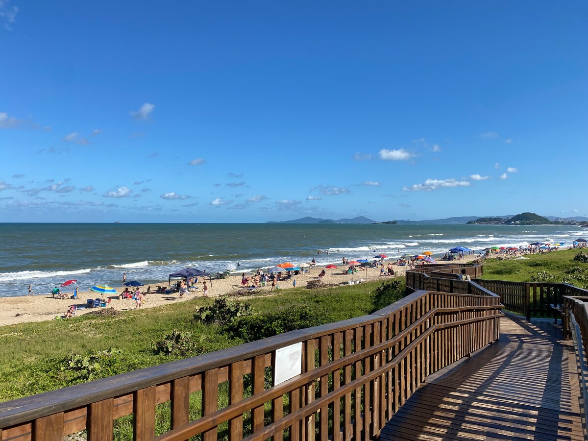 A scenic view showcases a sandy beach lined with colorful umbrellas and beachgoers. Waves can be seen gently rolling onto the shore, while a wooden boardwalk leads towards the beach, surrounded by lush greenery and the expansive ocean under a clear blue sky.