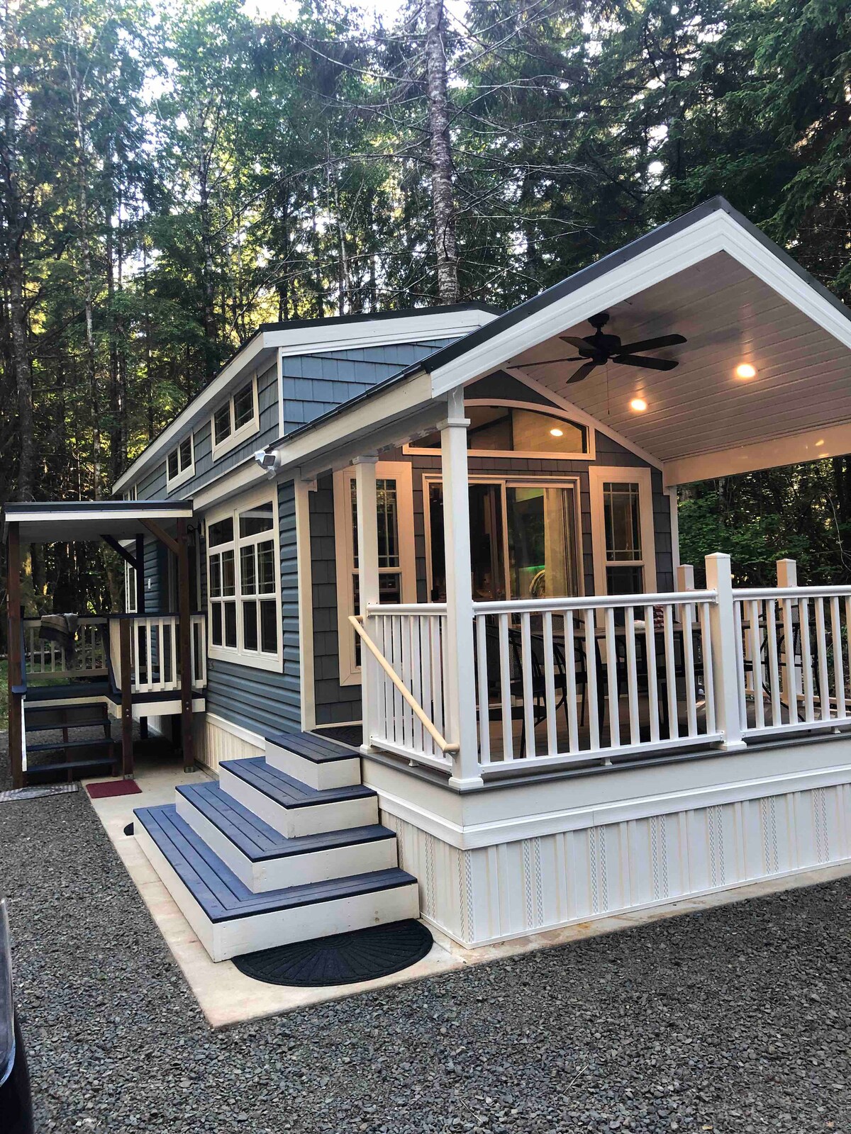 A charming cabin is set amidst tall trees, featuring a welcoming porch with a white railing and steps leading up to the entrance. Windows allow natural light to brighten the interior, enhancing the connection with the surrounding forest.
