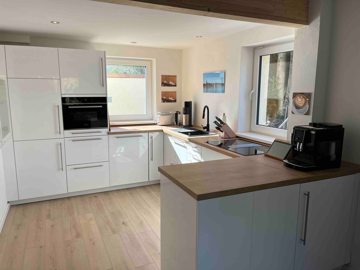 A modern kitchen is presented, featuring sleek white cabinetry and a wooden countertop. Under-cabinet lighting highlights the workspace. Appliances including an oven, microwave, and coffee machine are visible, alongside a sink with a stylish black tap. Natural light enters through a window with a serene outdoor view.