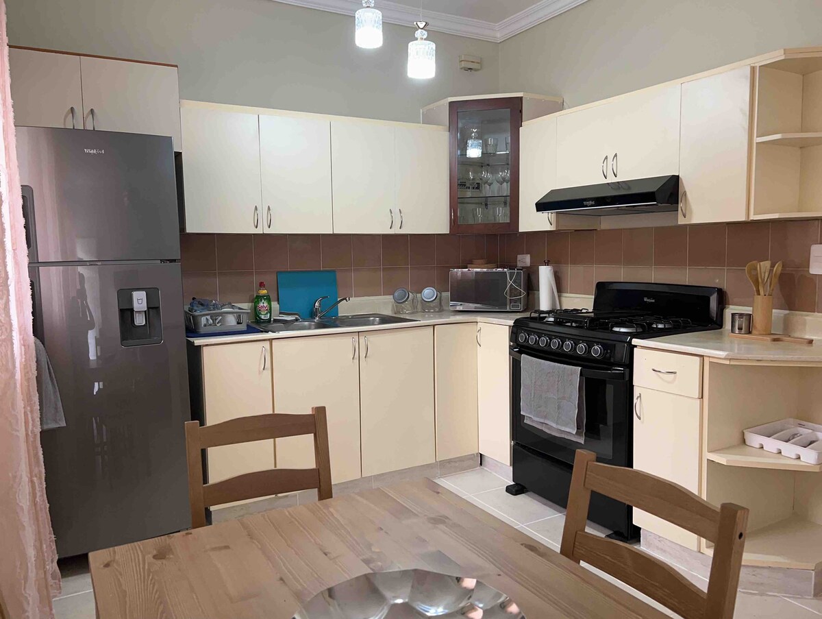 A well-equipped kitchen features modern appliances, including a stainless steel refrigerator and a gas stove. Cream-colored cabinets and countertops provide ample storage and workspace. A wooden dining table is positioned nearby, complemented by simple chairs, creating a functional and inviting dining area.