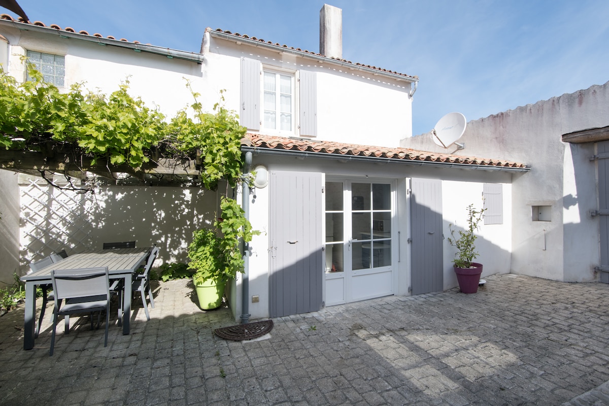 A charming exterior of a house is presented, featuring light grey shutters and a patio area. A dining table is arranged on the stone-paved courtyard, surrounded by climbing vines and potted plants. Sunlight illuminates the space, creating a bright and welcoming atmosphere.