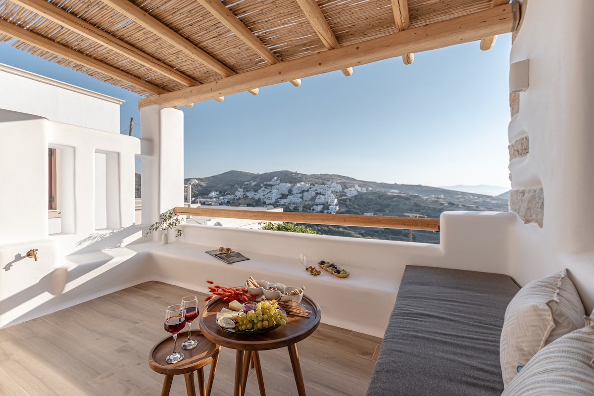 A spacious outdoor terrace features a wooden roof and a comfortable seating area. A wooden table is set with wine glasses and a variety of snacks. The serene landscape of the surrounding hills and village is visible in the background under a clear sky.