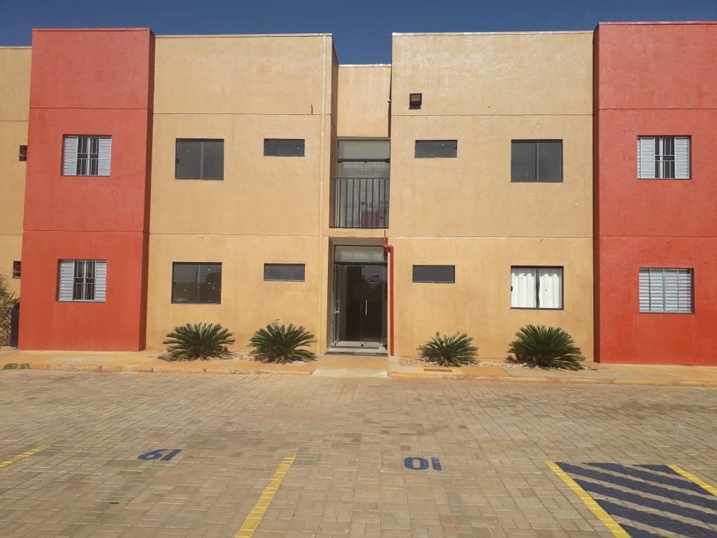 A two-story building is shown with a dual-tone exterior in orange and red. The façade is accented by rectangular windows and well-maintained landscaping featuring low shrubs. A paved parking area is visible in front, with designated spots marked on the surface.