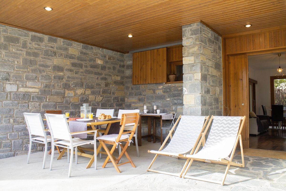 A shaded outdoor dining area features a wooden table set up with chairs, inviting guests to enjoy meals al fresco. Two reclining chairs are positioned for relaxation, while the stone walls and wooden beams create a rustic setting. Natural light enhances the inviting feel of the space.