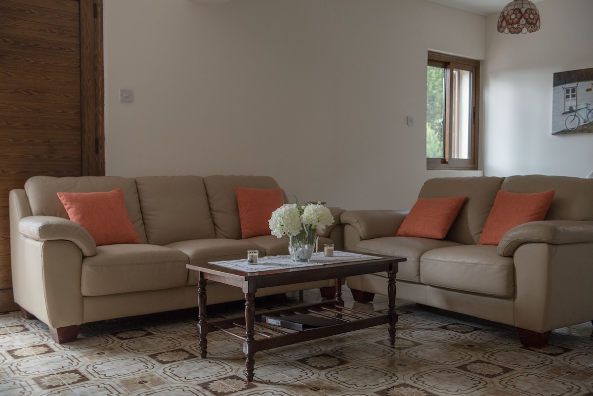 A comfortable living area features two cream-colored sofas adorned with orange cushions. A wooden coffee table sits in the center, displaying a vase of white flowers. Natural light enters through a nearby window, illuminating the space.
