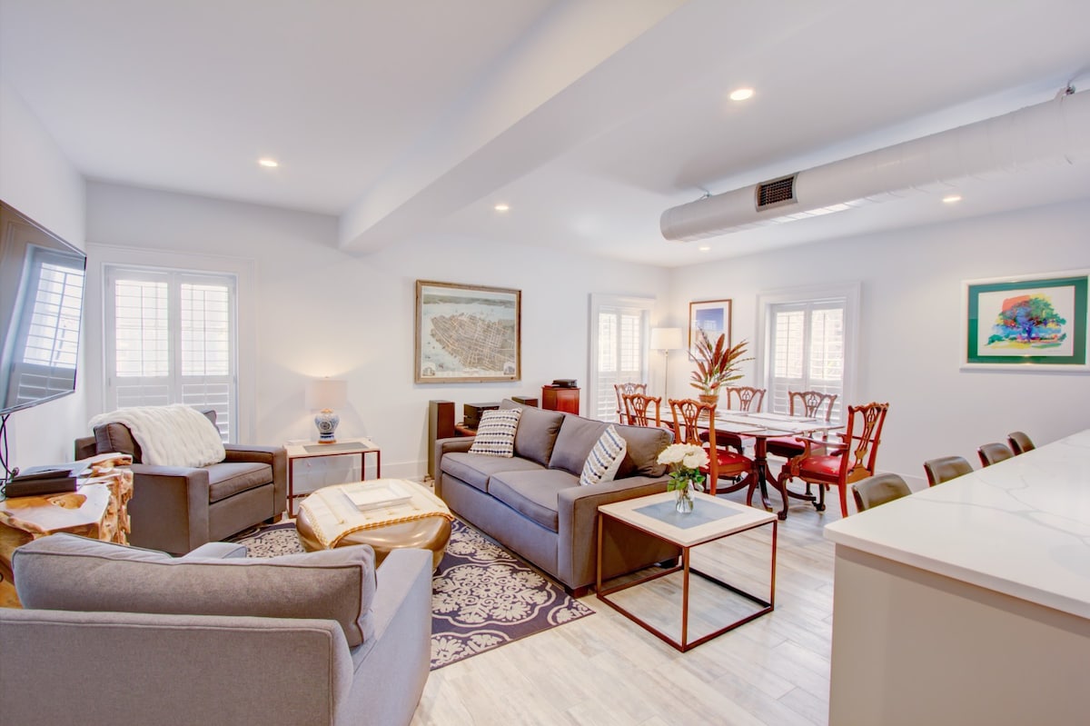 An open concept living area features a spacious sofa, two armchairs, and a coffee table on a patterned rug. A dining table with chairs is visible in the background, while light filters through white shutters on several windows, creating a bright and airy atmosphere.