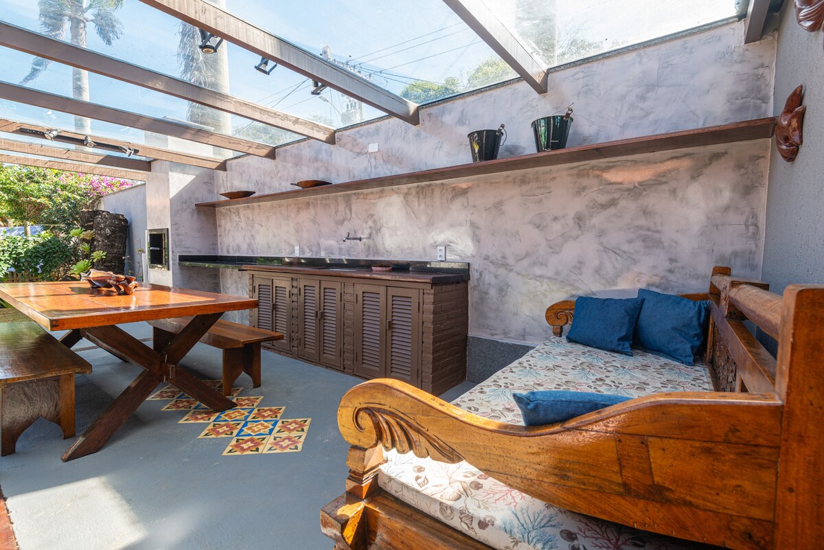 An outdoor kitchen area is highlighted by a wooden dining table and a comfortable seating space. The countertop features ample storage beneath, with decorative tiles adding color to the flooring. Clear roofing allows natural light to illuminate the space, surrounded by tropical plants.