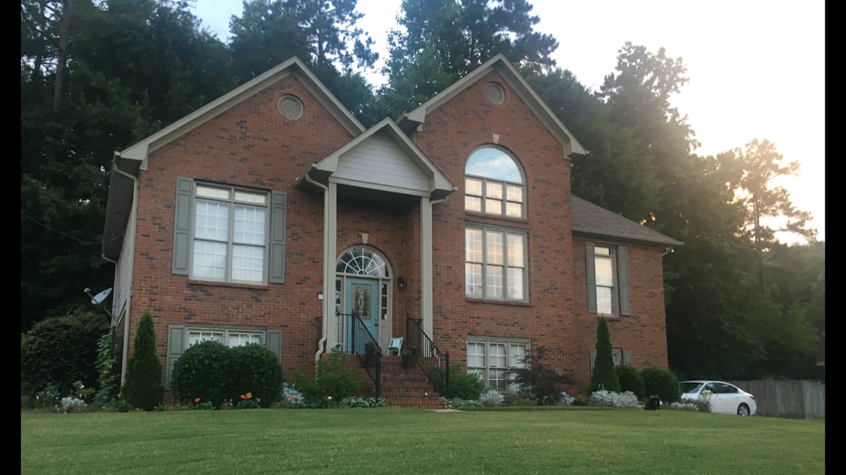 A spacious two-story brick house features large, arched windows framed by decorative shutters. The entryway is marked by a central door and flanked by steps leading to an inviting porch. Lush greenery and a manicured lawn surround the property, creating a welcoming exterior.