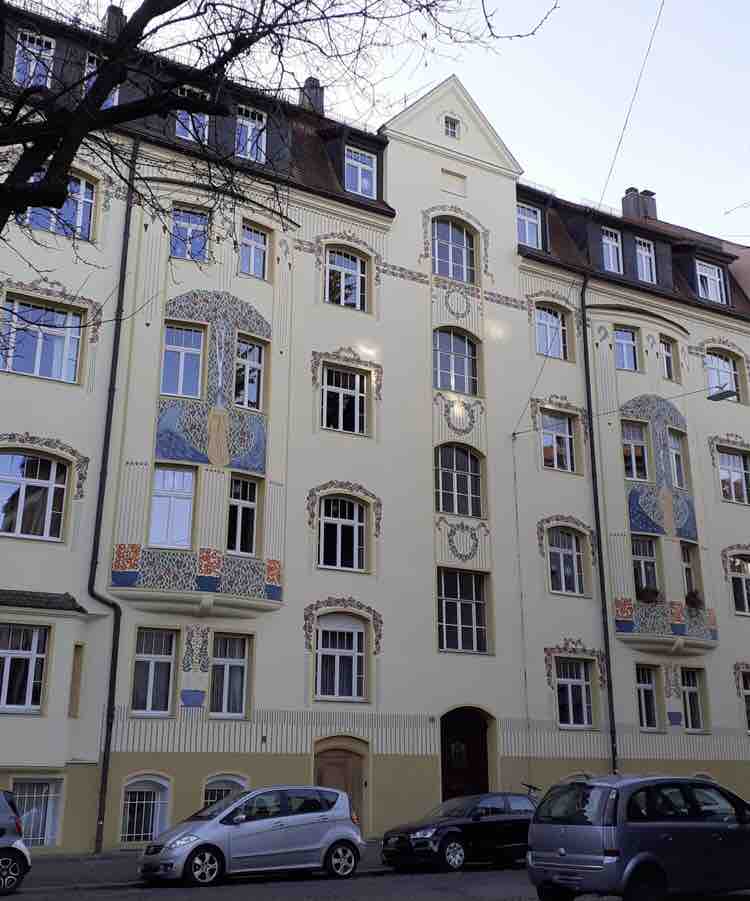 A charming apartment building features intricate decorative elements on its façade, blending traditional and modern architectural designs. Multiple windows are adorned with patterned details, allowing natural light to illuminate the interior. Cars are parked along the street, highlighting the urban setting.