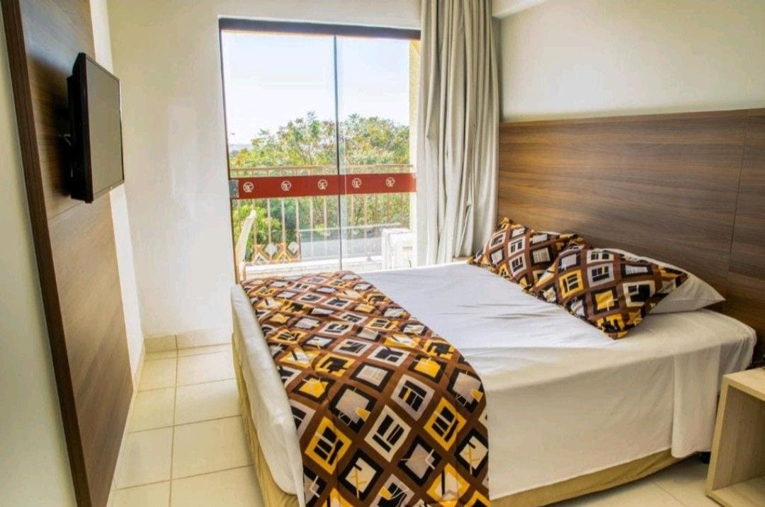 A well-appointed bedroom features a spacious bed adorned with a patterned blanket. The room is illuminated by natural light from a large window, which overlooks a balcony. A flat-screen TV is mounted on the wall, enhancing the comfort of the space.
