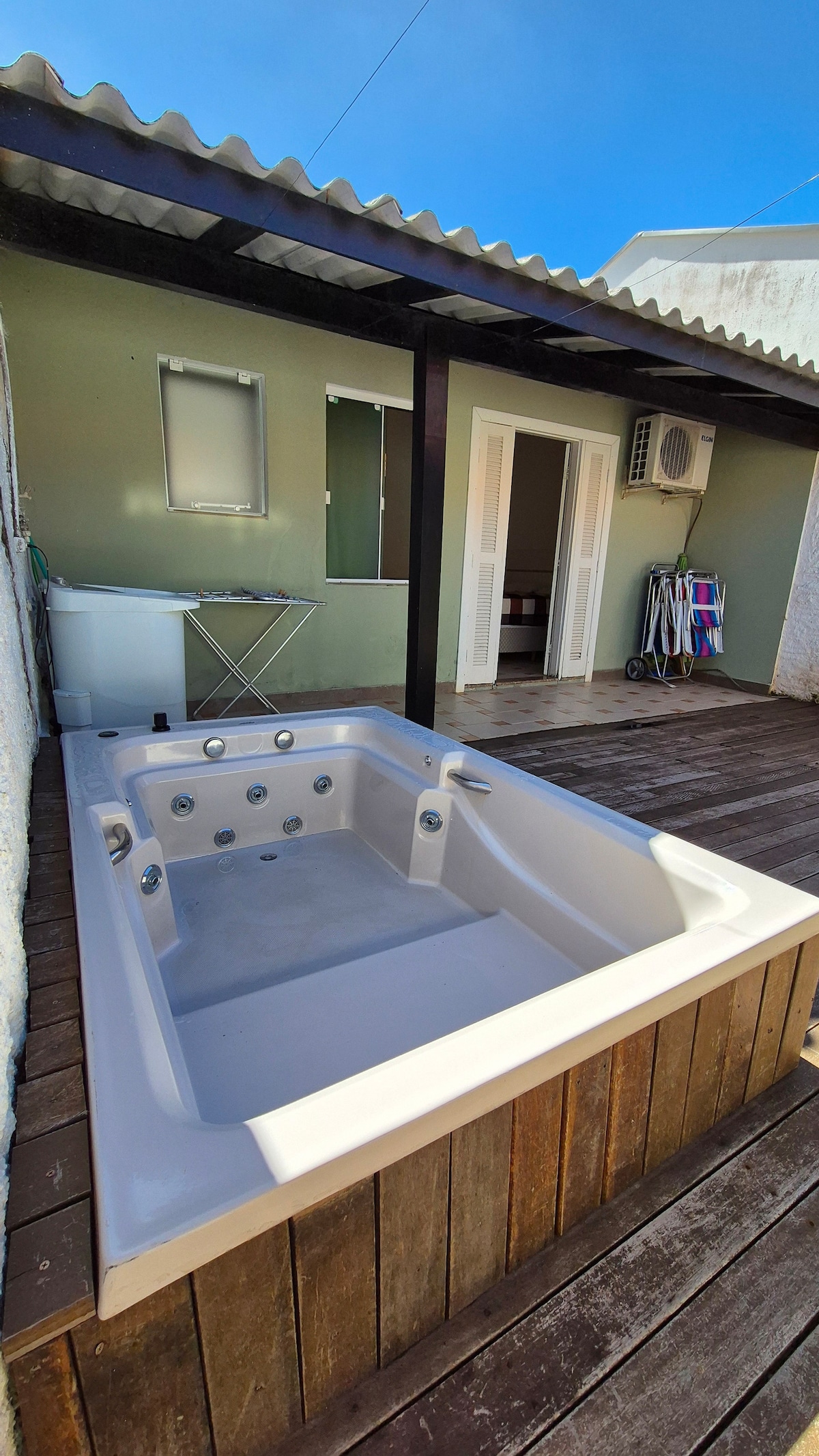 A spacious outdoor area features a wooden deck with a hot tub. The hot tub is surrounded by a simple wall and is adjacent to a building with a sliding glass door and windows. Nearby, a trellis supports a cover, providing shade.