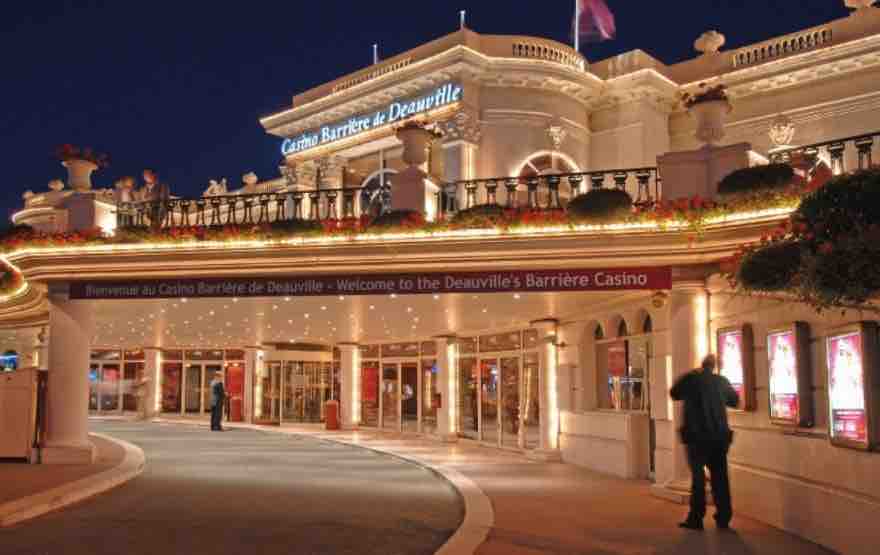 The exterior of Casino Barrière de Deauville is illuminated at night, with elegant architectural details and decorative planters lining the entrance. The welcoming signage invites guests to explore, while the rounded drive leads up to the grand entrance, enhancing the ambiance of this leisure destination.