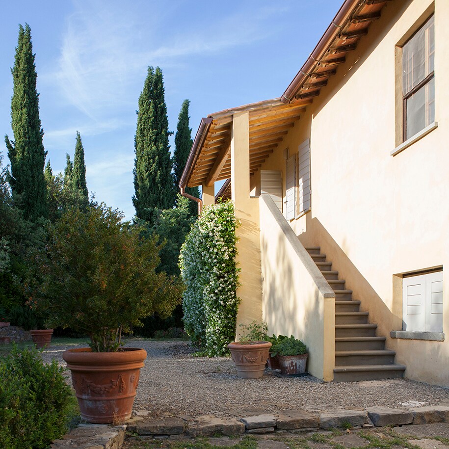 The exterior features a warm yellow facade with wooden roof beams. A set of stairs leads to the entrance, flanked by decorative pots and lush greenery. Tall cypress trees are visible in the background, contributing to a serene, landscaped setting.