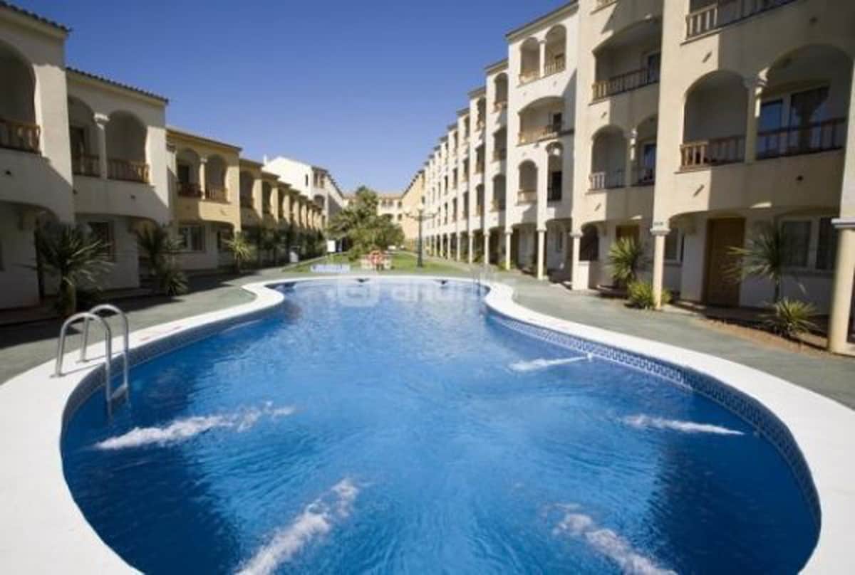 A large swimming pool is situated in the center of the complex, surrounded by several multi-story buildings. The water is a clear blue, with gentle ripples visible. Tall palm plants line the edges, providing a green contrast against the architecture and the clear sky.
