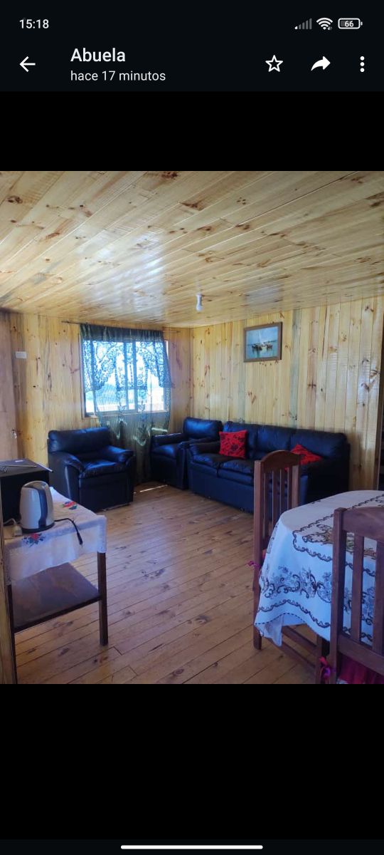 A cozy living area features dark leather sofas, complemented by decorative pillows. Natural wood paneling and flooring create a warm ambiance. A small wooden dining table is visible, with a white tablecloth, and a kettle sits on the nearby surface. A window allows natural light to enter.