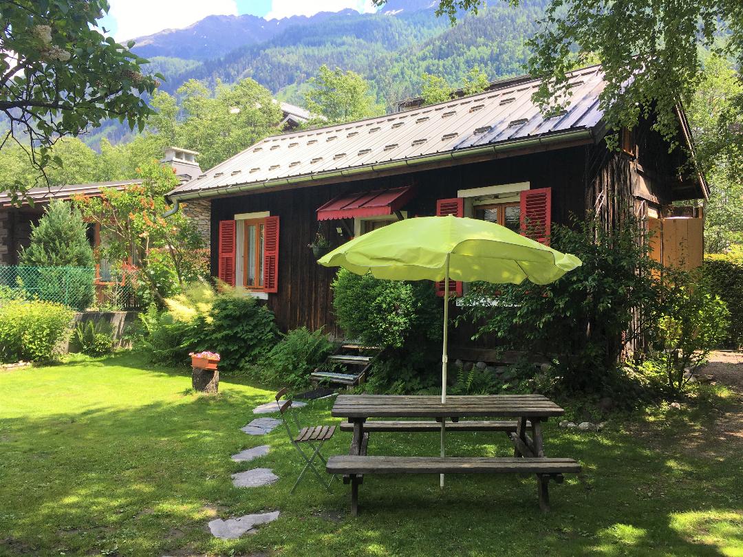 A charming independent chalet is set amidst greenery, featuring vibrant red shutters on its wooden façade. A picnic table with an umbrella provides a shaded outdoor seating area on the well-kept lawn, accented by stone pathways and surrounding trees.
