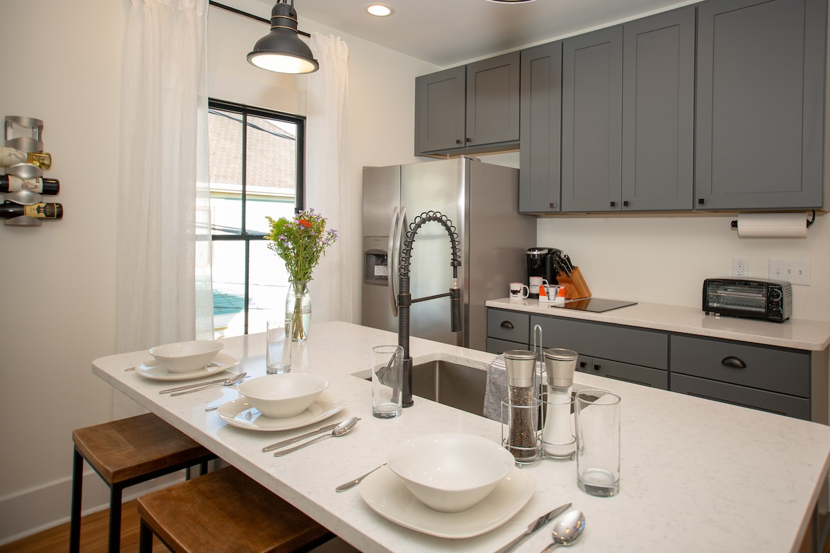 A modern kitchen features gray cabinetry and a large central island with seating for four. A stainless steel refrigerator and various kitchen appliances are present. Natural light fills the space through a window, while a vase of fresh flowers adds a touch of color.