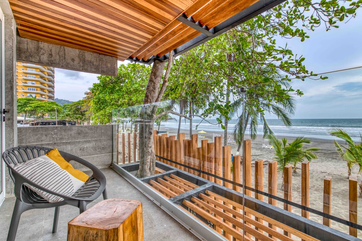 A serene terrace features a sleek black chair paired with a wooden stool, offering a perfect spot to relax. Large glass panels provide unobstructed ocean views, while lush greenery and palm trees enhance the beachfront setting.