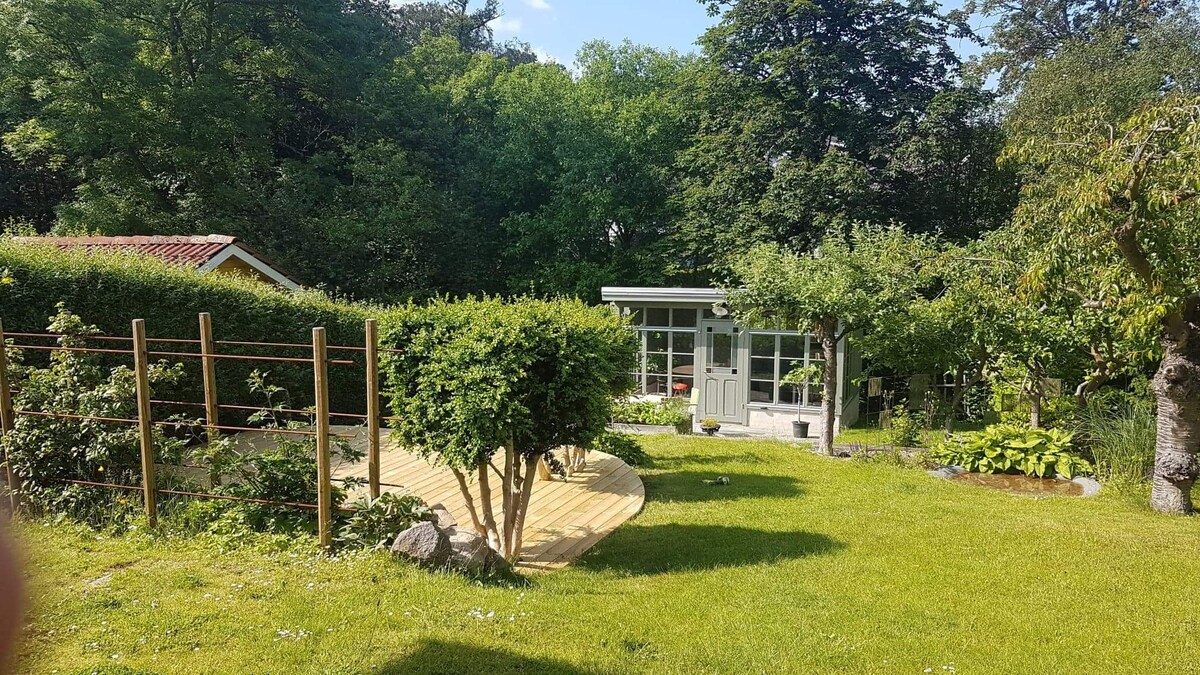 An inviting garden space features a well-maintained lawn surrounded by lush greenery and trees. A wooden deck leads towards a charming glass structure, which serves as a cozy retreat. Neatly arranged shrubs and a variety of plants add to the tranquil environment.