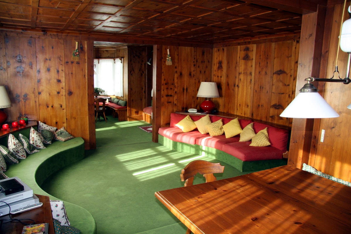 A cozy living area features wooden paneling and green carpet. Curved seating with red cushions is arranged next to a wooden table. Two table lamps provide soft lighting, while natural light filters through the window, creating a warm ambiance.