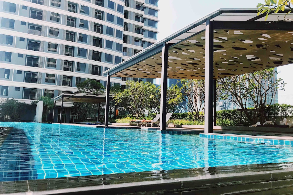 A serene swimming pool is featured, with turquoise tiles reflecting sunlight. Shaded lounge areas with modern canopies are present beside the pool, surrounded by landscaped gardens. The impressive building structure provides a backdrop, creating a relaxing atmosphere for guests.