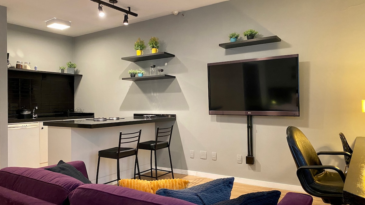 An open-plan space features a modern kitchen with sleek black countertops and shelves adorned with small potted plants. A large flat-screen TV is mounted on the wall. Comfortable seating is provided with a purple sofa and a black office chair positioned nearby.