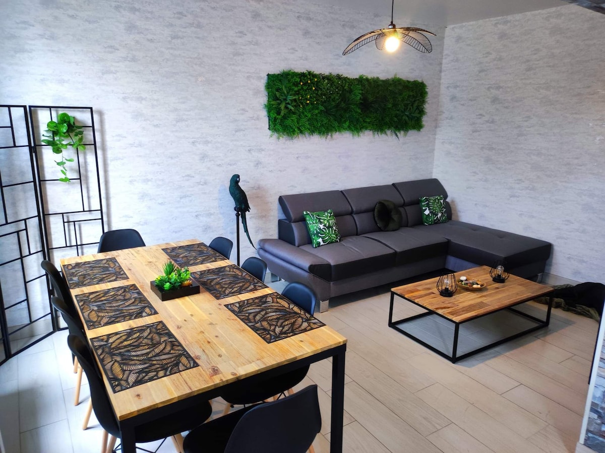 A modern living area features a comfortable sectional sofa adorned with green accents. A wooden coffee table is positioned in front, with decorative items arranged on top. A dining table with black chairs is nearby, complemented by wall art and a vertical garden element.