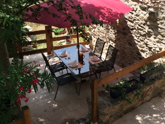A shaded dining area is positioned under a large umbrella, featuring a glass-top table surrounded by six metal chairs. Lush greenery and flower pots add a touch of color to the stone walls, creating a serene outdoor space for meals.