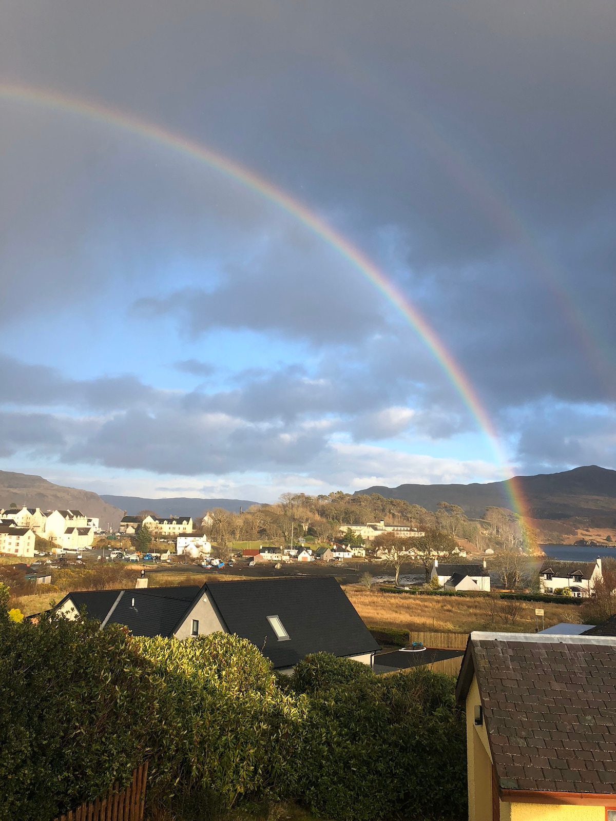 A vibrant rainbow arcs over the town, with rooftops of houses visible below. The scene features lush greenery and mountains in the background, set against a partially cloudy sky that allows sunlight to filter through.