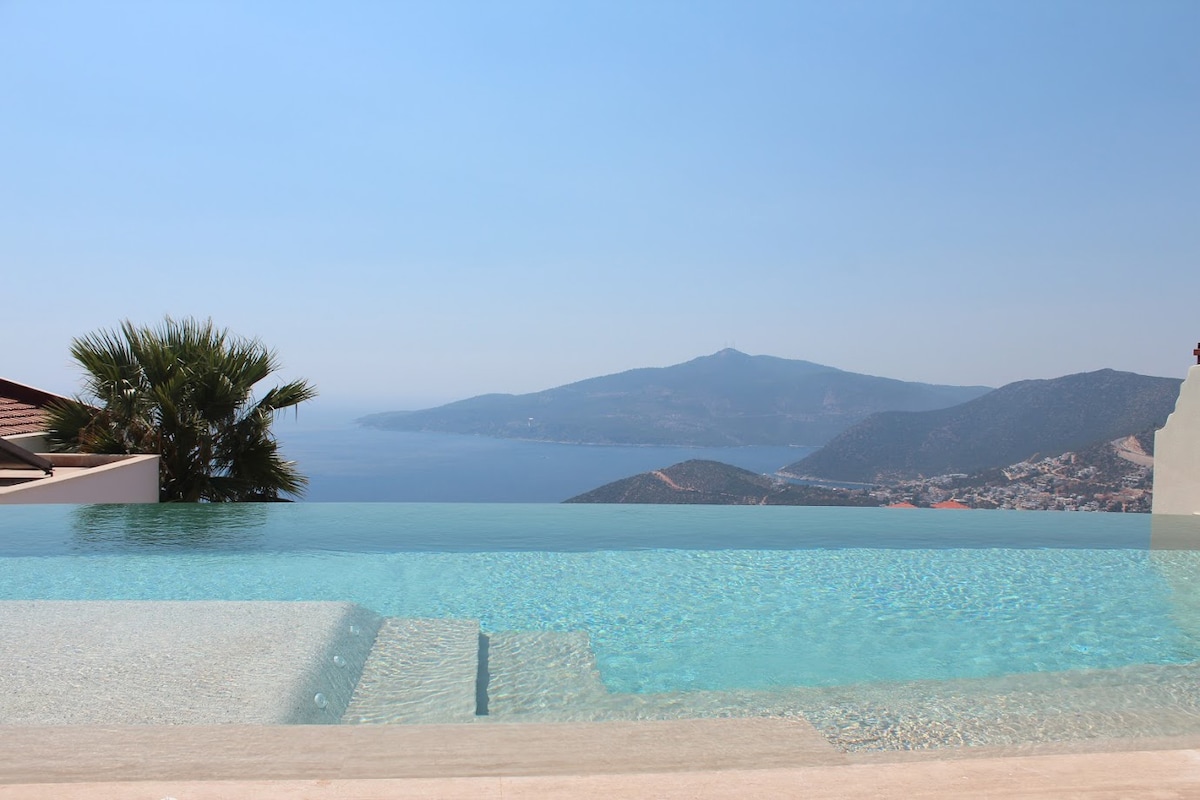 An infinity pool stretches towards the horizon, showcasing a seamless transition between water and sea with gentle blue hues. The surrounding landscape features mountainous terrain, adding depth to the scenic vista. Clear skies complement the tranquil ambiance, perfect for relaxation.