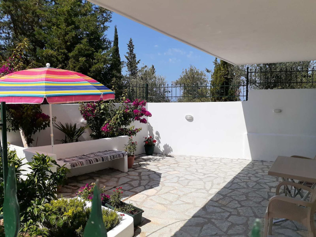 A spacious outdoor yard is presented, featuring a striped umbrella providing shade over a seating area. Colorful flowers and greenery enhance the natural setting, while a stone-paved space allows for relaxation or dining. The atmosphere reflects a peaceful retreat in a quiet environment.