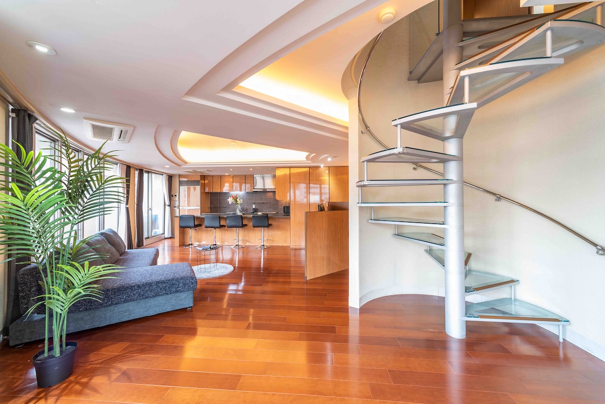 A spacious living area is highlighted by glossy wooden flooring and modern spiral staircase. Large windows invite natural light, enhancing the open layout. A dark sofa and round rug provide comfortable seating. The kitchen area, visible in the background, features sleek cabinetry and bar seating.