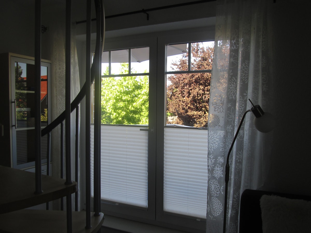 A light-filled living area showcases large glass doors leading to a balcony. Sheer white curtains add softness, while plants outside bring a touch of greenery. A spiral staircase is positioned nearby, contributing to the open layout of the space.