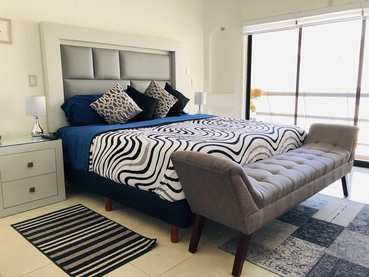 A spacious bedroom features a large bed dressed in a bold black-and-white striped comforter. Two decorative pillows add texture, while a sleek bench sits at the foot of the bed. Soft light enters through large glass doors, enhancing the calm atmosphere.