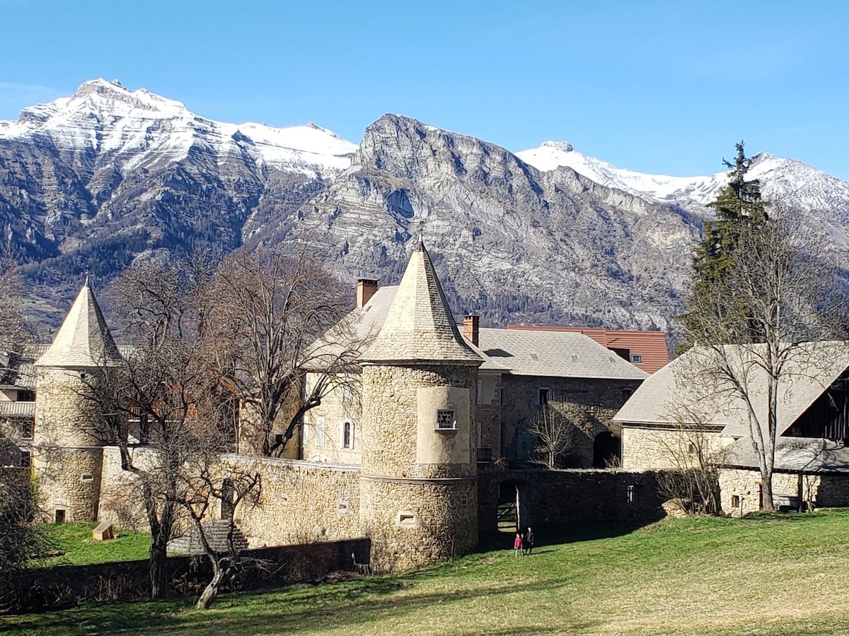A scenic view showcases a stone building with distinctive tower features set against a backdrop of snow-capped mountains. A grassy area lies in the foreground, while two figures are seen walking along the path, contributing to the tranquil outdoor setting.