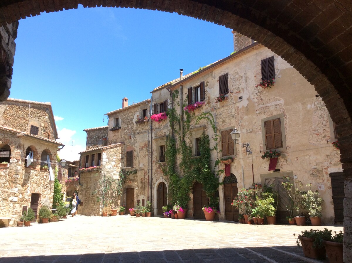 An archway frames a quaint courtyard with stone buildings adorned in climbing greenery and vibrant flowers. Terracotta pots are arranged along the edges, adding color. A clear blue sky enhances the warm ambiance of this historical setting, providing a glimpse into the village life.