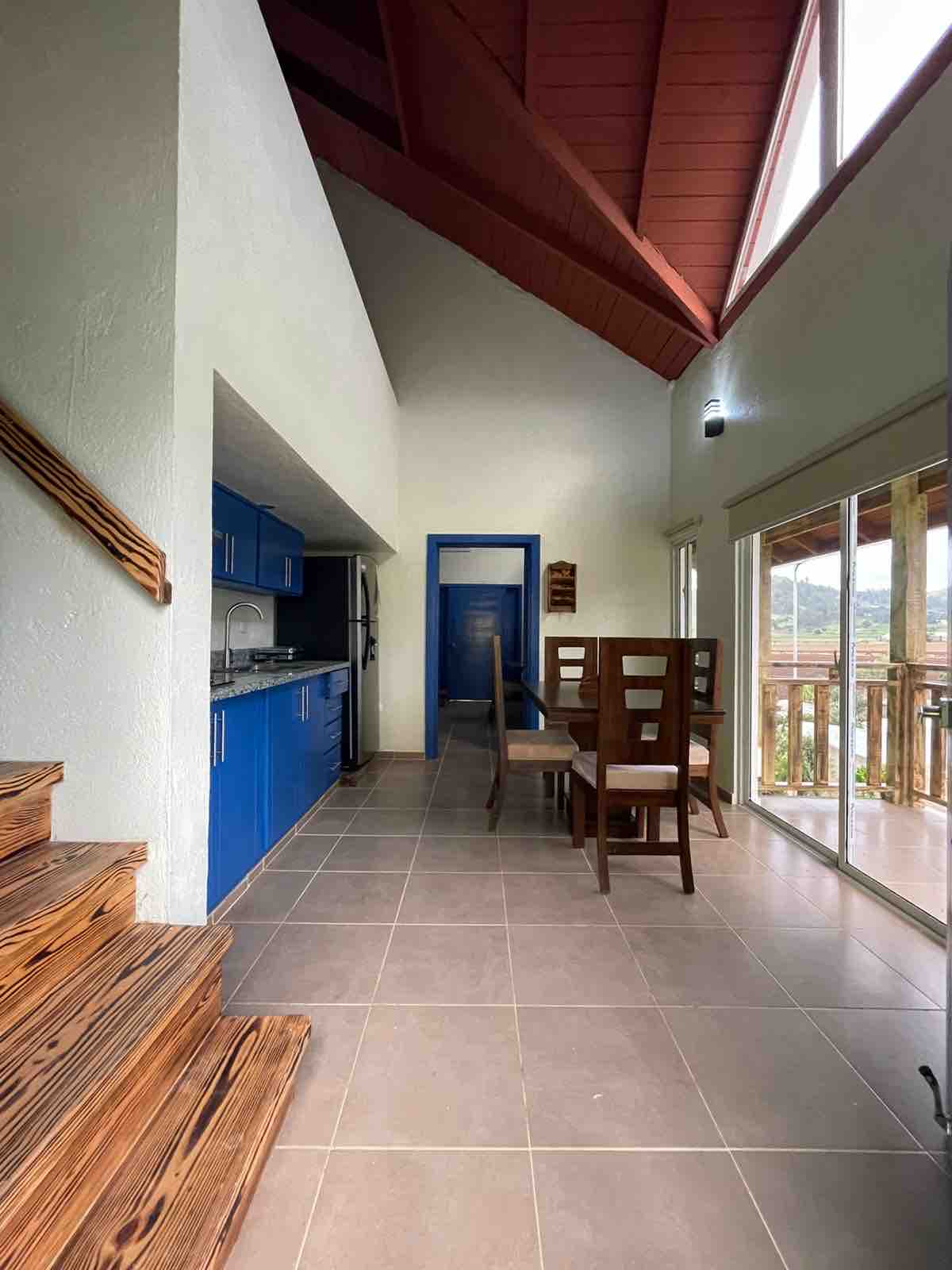 A spacious dining area is located adjacent to the kitchenette, featuring a wooden table surrounded by four chairs. Large windows provide natural light and showcase views of the balcony and surrounding landscape. The high ceiling emphasizes the open layout, with wooden stairs visible leading to the upper level.