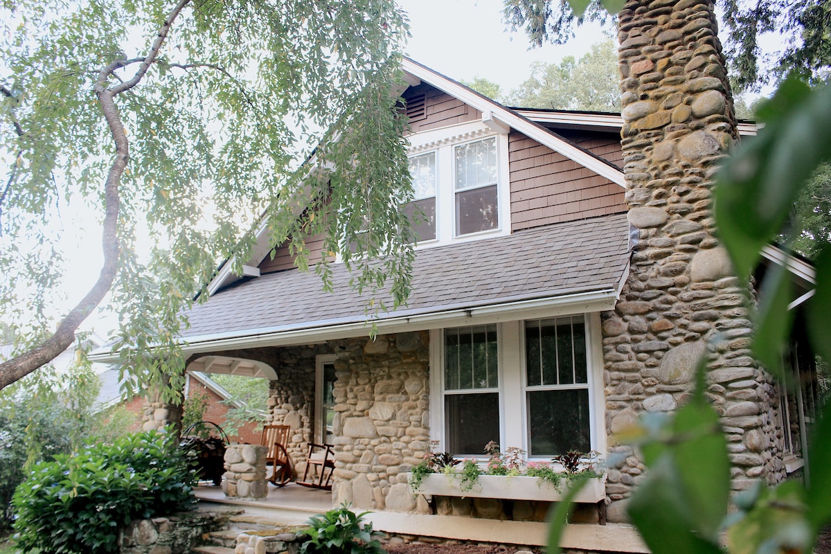 The cottage's exterior reveals a blend of stone and wood detailing. A welcoming front porch holds rocking chairs, surrounded by leafy greenery. Natural light illuminates the windows, which enhance the home’s traditional charm.